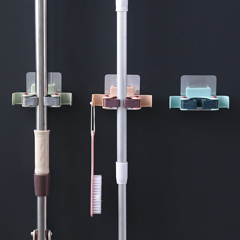 Green, pink, and light blue adhesive wall clips holding cleaning handles, with side hooks for lightweight tools